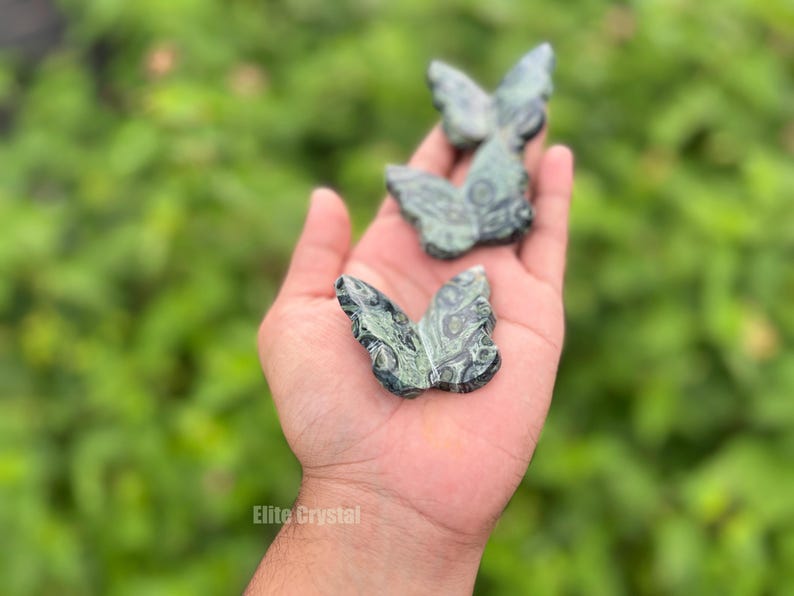 May include: Three green and black butterfly figurines are held in an open hand. The butterfly sculptures have detailed wing patterns. The background is a blurred green. The text "Elite Crystal" is visible at the bottom.