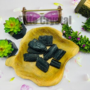 May include: A collection of raw, black tourmaline crystals displayed in a wooden bowl. The crystals have a rough, textured surface. The arrangement is complemented by decorative elements, including a miniature hourglass and artificial succulents.