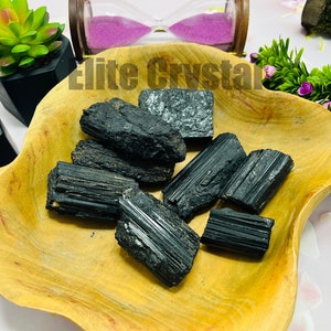 May include: A collection of raw, black tourmaline crystals displayed in a wooden bowl. The crystals vary in size and shape, with visible striations and a natural, unpolished appearance. The bowl sits on a white surface, with decorative plants and an hourglass in the background.