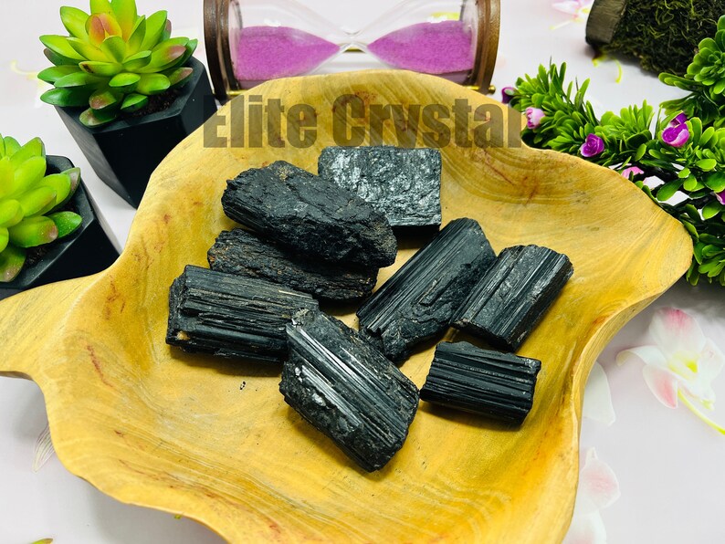 May include: A collection of raw, black tourmaline crystals displayed in a wooden bowl. The crystals vary in size and shape, with visible striations and a natural, unpolished appearance. The bowl sits on a white surface, with decorative plants and an hourglass in the background.