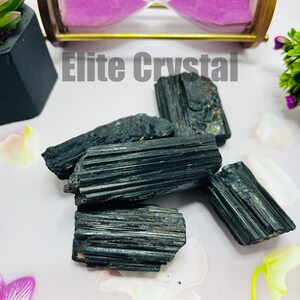 May include: A collection of raw, black tourmaline crystals. The crystals have a striated texture and are arranged on a light pink surface, with the text "Elite Crystal" in the background. Decorative plants and an hourglass are also present.