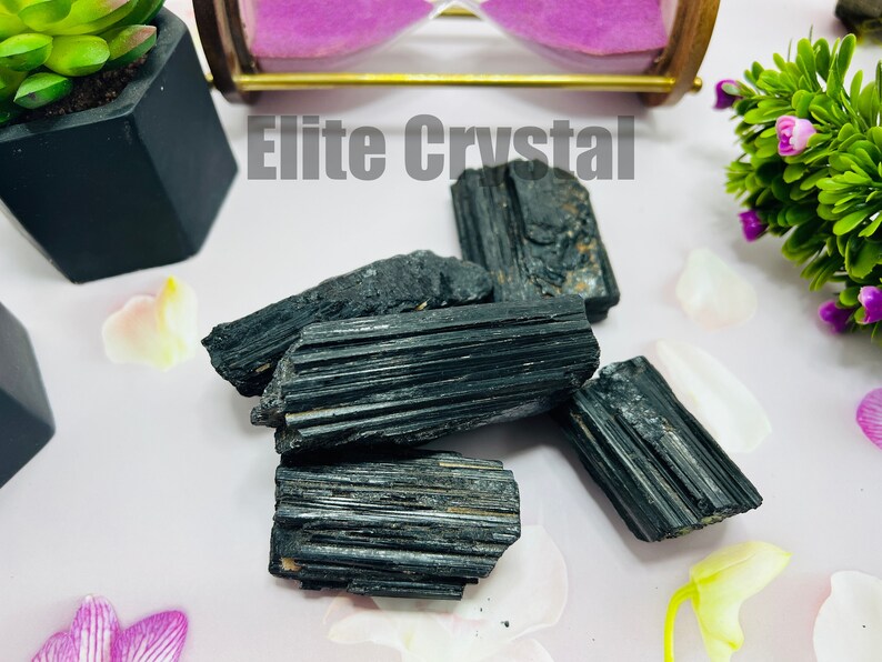 May include: A collection of raw, black tourmaline crystals. The crystals have a striated texture and are arranged on a light pink surface, with the text "Elite Crystal" in the background. Decorative plants and an hourglass are also present.