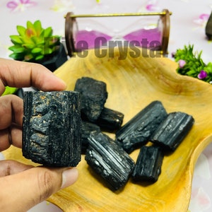 May include: A hand holding a black tourmaline crystal, with several other raw black tourmaline stones in a wooden bowl. The crystals have a rough, textured surface. The background includes a small hourglass and greenery.