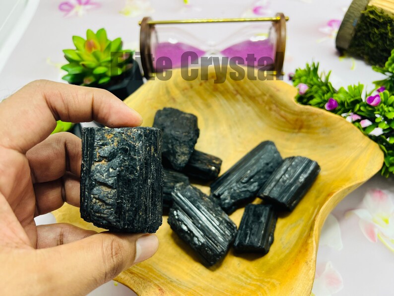 May include: A hand holding a black tourmaline crystal, with several other raw black tourmaline stones in a wooden bowl. The crystals have a rough, textured surface. The background includes a small hourglass and greenery.