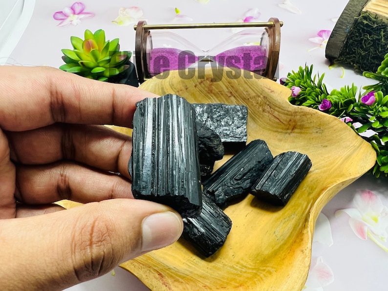 May include: A collection of raw, black tourmaline crystals displayed in a wooden bowl. The crystals have a striated texture and varying sizes. The bowl is set against a background with decorative elements, including a small hourglass and greenery.