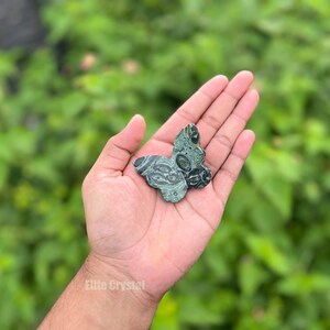 May include: A green and black butterfly-shaped stone carving. The stone has a patterned design and is held in an open hand against a blurred green background. The stone carving is approximately 2 inches wide.