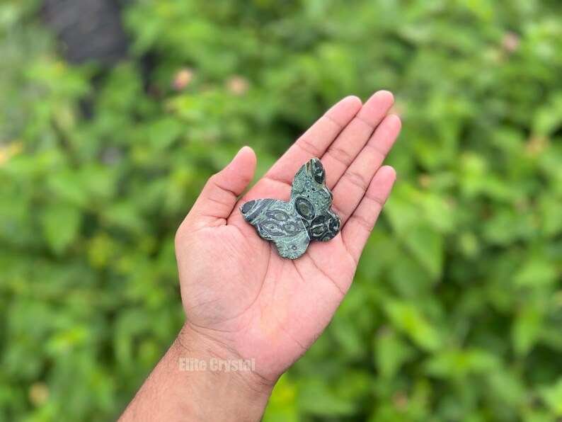 May include: A green and black butterfly-shaped stone carving. The stone has a patterned design and is held in an open hand against a blurred green background. The stone carving is approximately 2 inches wide.