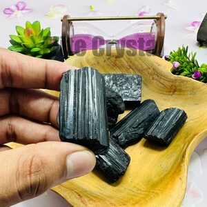 May include: A collection of raw, black tourmaline crystals displayed in a wooden bowl. The crystals have a striated texture and varying sizes. The bowl is set against a background with decorative elements, including a small hourglass and greenery.