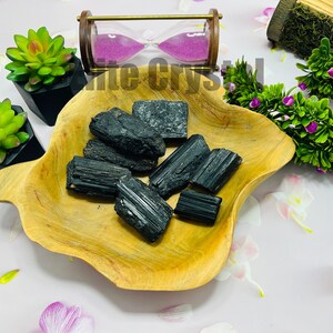 May include: A collection of raw, black tourmaline crystals displayed in a light brown, wooden, leaf-shaped bowl. The crystals have a rough, textured surface. Decorative succulents and an hourglass are in the background.