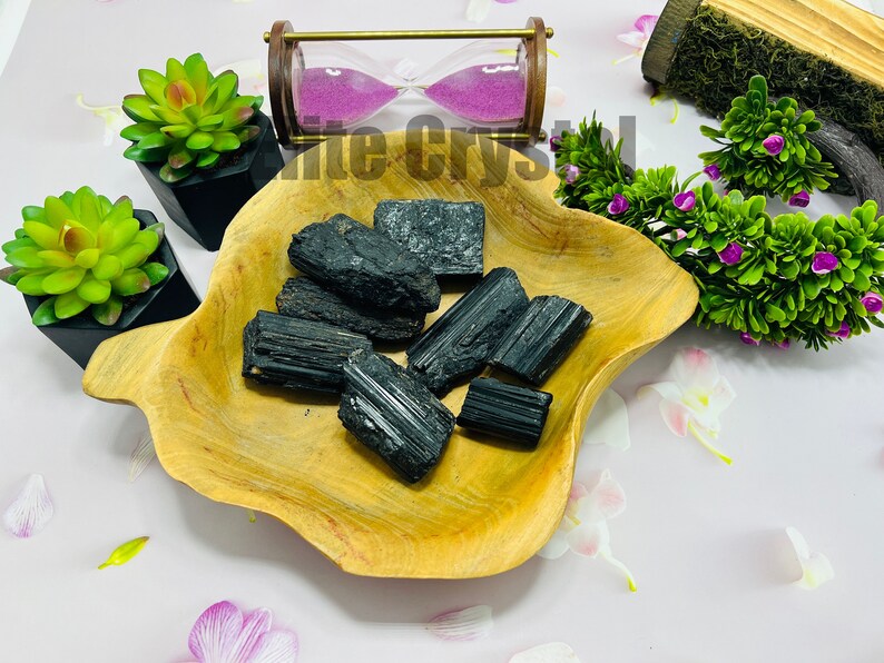 May include: A collection of raw, black tourmaline crystals displayed in a light brown, wooden, leaf-shaped bowl. The crystals have a rough, textured surface. Decorative succulents and an hourglass are in the background.