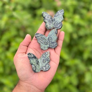 May include: Three green and black butterfly figurines are held in a hand against a blurred green background. The butterfly sculptures have a detailed pattern and are made of a stone-like material. The butterflies are a decorative item.