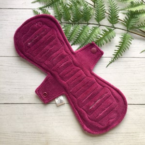 Puede incluir: Primer plano de una compresa menstrual reutilizable de tela rosa oscuro con una superficie texturizada y dos cierres a presión. La compresa tiene una etiqueta blanca con las letras "S/G" impresas en ella.