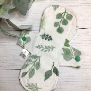 Peut inclure: Un protège-slip en tissu réutilisable blanc avec un motif de feuilles vertes. Le protège-slip a une fermeture à pression et est étiqueté "S/G".