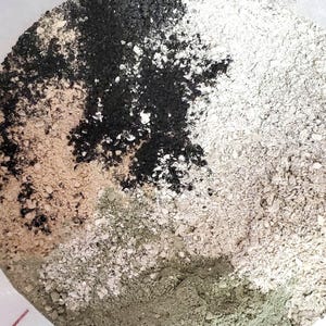 May include: A circular container filled with a mixture of loose, dry powders in distinct sections. The composition includes patches of fine black charcoal, light beige sand, coarse off-white granules, and a smooth, muted olive green powder.