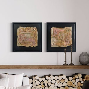 May include: Two framed abstract art pieces with gold and pink textured designs, displayed on a wooden shelf above a stacked wood log wall, with a black leather armchair and a vase.