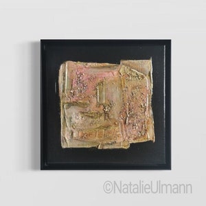 May include: Abstract painting with gold and pink textured layers in a black frame. The painting has a rough, textured surface and is framed in a black frame.