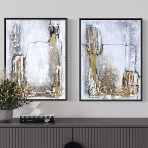 May include: Two abstract paintings in black frames with a white, gray, and gold color scheme. The paintings are hung on a white wall above a gray dresser.
