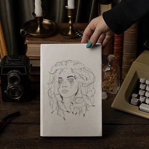 May include: A white sketchbook with a pencil drawing of a woman with snakes in her hair. The drawing is on a wooden table with a vintage camera, a framed print of mushrooms, and a typewriter.