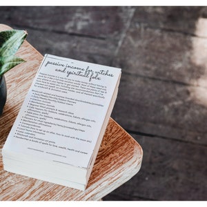 May include: A stack of white papers with black text titled "Passive Income for Witches and Spiritual Folk". The text lists ideas for passive income for witches, such as creating printable planners, journals, and spell books.