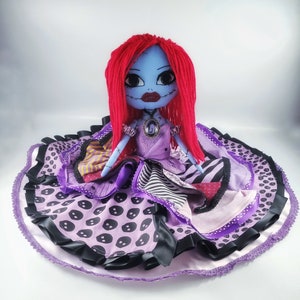 May include: A handmade doll with red yarn hair, blue skin, and a purple and black patchwork dress. The doll is sitting on a purple and black fabric with a white lace trim.