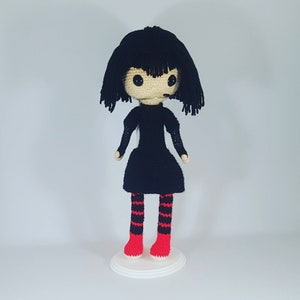 May include: A crocheted doll with black hair, a black dress, and red and white striped stockings. The doll is standing on a white stand.