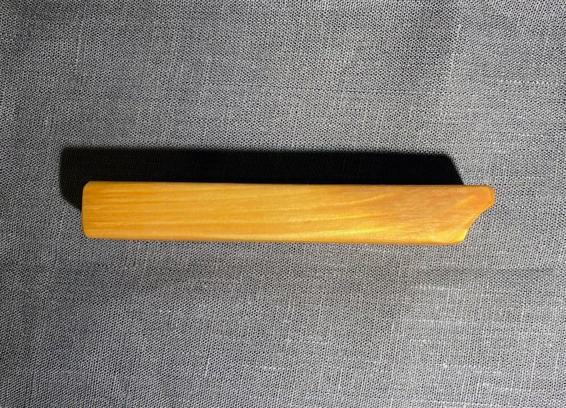 Puede incluir: Una pinza para el cabello rectangular de madera con un acabado liso de color marr&oacute;n claro y un extremo inclinado y c&oacute;nico. El accesorio presenta un patr&oacute;n de veta de madera natural en toda su superficie, con un dise&ntilde;o simple, alargado y plano.