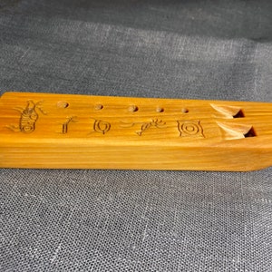 Puede incluir: Una flauta rectangular de madera marr&oacute;n claro con cinco orificios circulares para los dedos y dos aberturas sonoras talladas. La superficie est&aacute; decorada con seis s&iacute;mbolos grabados distintos, que incluyen una espiral, una figura estilizada y patrones geom&eacute;tricos.
