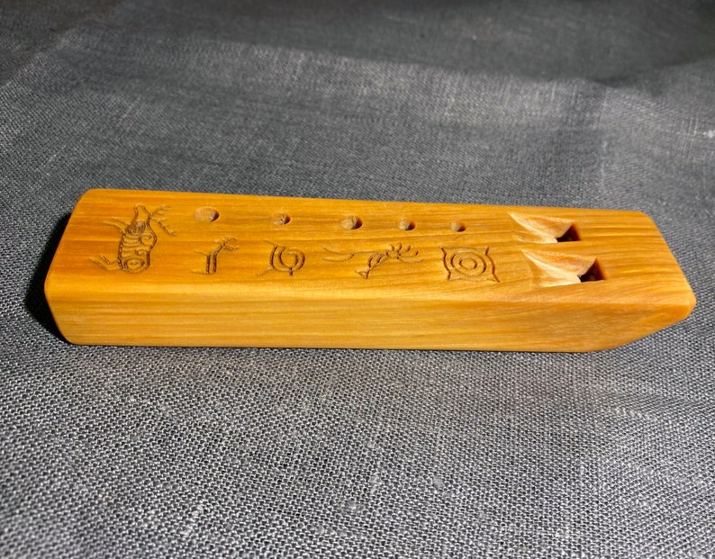 Puede incluir: Una flauta rectangular de madera marr&oacute;n claro con cinco orificios circulares para los dedos y dos aberturas sonoras talladas. La superficie est&aacute; decorada con seis s&iacute;mbolos grabados distintos, que incluyen una espiral, una figura estilizada y patrones geom&eacute;tricos.