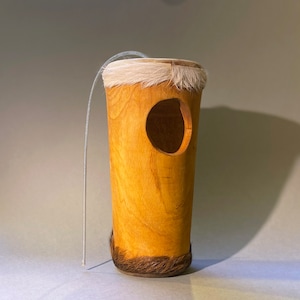 Puede incluir: Un instrumento de percusión de madera con un acabado natural. El tambor tiene una abertura redonda en el lateral y está cubierto con una piel de animal blanca. Una cuerda fina está unida a la parte superior del tambor.