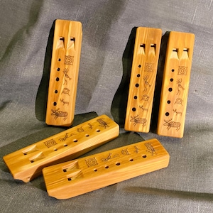 Puede incluir: Cuatro flautas rectangulares de madera con veta marr&oacute;n claro, cada una con dos orificios de aire en la parte superior, una fila de orificios para los dedos y s&iacute;mbolos grabados con l&aacute;ser que incluyen una figura, un ciervo y c&iacute;rculos.