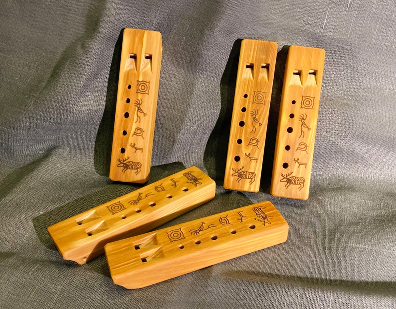 Puede incluir: Cuatro flautas rectangulares de madera con veta marr&oacute;n claro, cada una con dos orificios de aire en la parte superior, una fila de orificios para los dedos y s&iacute;mbolos grabados con l&aacute;ser que incluyen una figura, un ciervo y c&iacute;rculos.