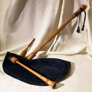 May include: A wooden bagpipe with a black leather bag and three pipes. The pipes are made of wood and have a brown finish. The bagpipe is lying on a white cloth background.