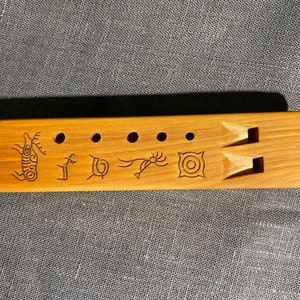 Puede incluir: Una flauta rectangular de madera marr&oacute;n claro con cinco orificios circulares y dos boquillas talladas. La superficie est&aacute; decorada con cinco s&iacute;mbolos distintos grabados en marr&oacute;n oscuro, incluyendo un ciervo, una espiral y un patr&oacute;n geom&eacute;trico.