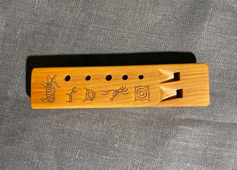 Puede incluir: Una flauta rectangular de madera marr&oacute;n claro con cinco orificios circulares y dos boquillas talladas. La superficie est&aacute; decorada con cinco s&iacute;mbolos distintos grabados en marr&oacute;n oscuro, incluyendo un ciervo, una espiral y un patr&oacute;n geom&eacute;trico.