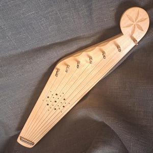 Puede incluir: Una lira de madera clara, con cuerdas y clavijas de afinación. El instrumento tiene una cabeza redondeada con un diseño floral tallado. La lira se encuentra sobre un fondo de tela gris oscuro, mostrando su veta natural y artesanía.