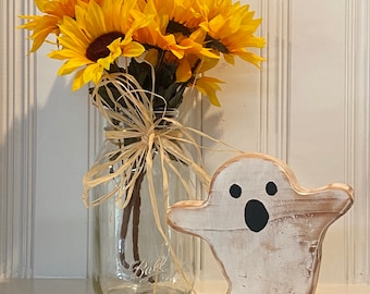 May include: A white wooden ghost with black eyes and a mouth, standing on a shelf next to a glass jar filled with yellow sunflowers.