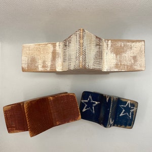 May include: Three wooden stars in the shape of a traditional American flag. The top star is whitewashed wood, the middle star is red, and the bottom star is blue with white stars.