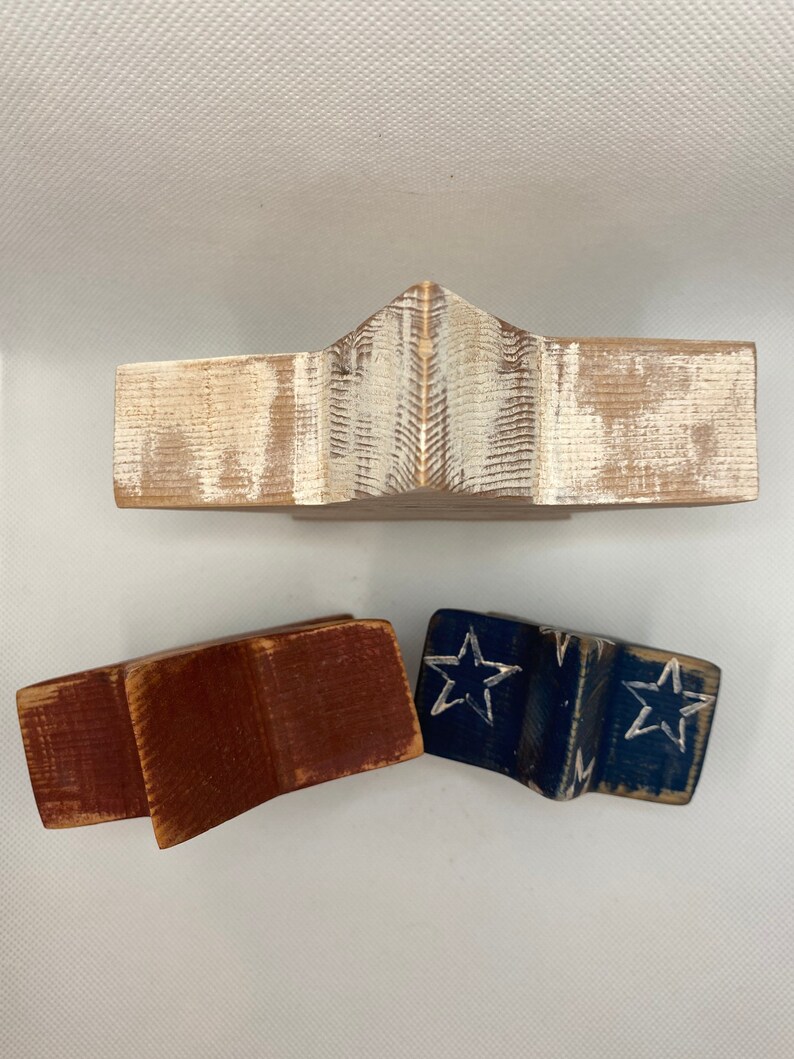 May include: Three wooden stars in the shape of a traditional American flag. The top star is whitewashed wood, the middle star is red, and the bottom star is blue with white stars.