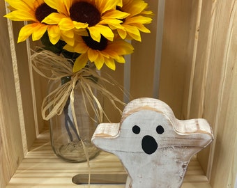 May include: A whitewashed wooden ghost figurine with black eyes and a mouth, sitting in a wooden crate with a bouquet of yellow sunflowers in a glass jar.