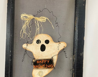 May include: A black wooden frame with a screen mesh background. A white ghost with black eyes and a bow is attached to the screen. A small wooden sign with the text "Happy Haunting" is attached to the ghost.