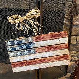 May include: A rustic wooden American flag with red, white, and blue paint. The flag is distressed and has a weathered look. It is hanging from a wire and has a raffia bow on top.