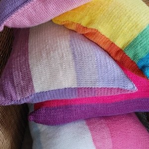 Könnte beinhalten: Ein Stapel bunter gestreifter Strickkissen in den Farben Rosa, Lila, Weiß, Gelb, Orange und Grün.