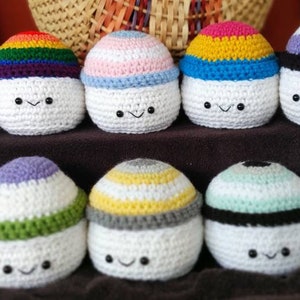 May include: Twelve crocheted plush eggs, each with a different color pattern representing a pride flag. The eggs are white with a crocheted design on top. Each egg has a smiling face.