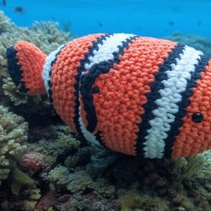 Pode incluir: Um peixe-palhaço de crochê com listras laranja, brancas e pretas, em um cenário de corais e água azul. O peixe feito à mão tem um corpo redondo e um olho de botão preto, mostrando um tema subaquático caprichoso.