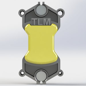 Może przedstawiać: Szary i żółty element mechaniczny z literami "TLM" na górze. Element ma centralną żółtą sekcję i szarą ramę zewnętrzną z okrągłymi otworami i zaokrąglonymi końcami.