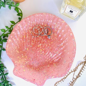May include: A pink seashell-shaped trinket dish with gold flecks, holding silver rings.