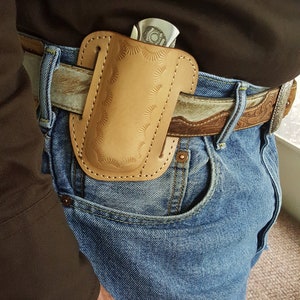 May include: A brown leather knife sheath with a decorative pattern is attached to a brown leather belt with a silver buckle. The belt is worn over a pair of blue jeans.