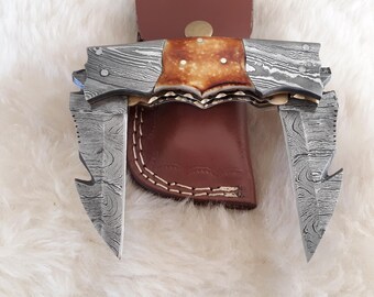 May include: A custom-made knife with a Damascus steel blade and a leather sheath. The knife features a unique design with a patterned blade and a brown leather handle. The sheath is also brown and has white stitching.