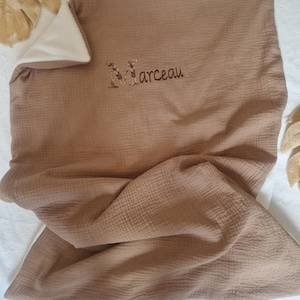 May include: A tan baby blanket with a white interior. The blanket has a crinkled texture and the name "Marceau" is embroidered in brown thread. The blanket is folded and lies on a white surface.