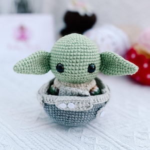 May include: A crocheted figure of a character with large ears and black eyes, seated in a gray and beige crocheted pod. The figure is light green, with a beige body and large, pointed ears. The pod is gray with white accents.
